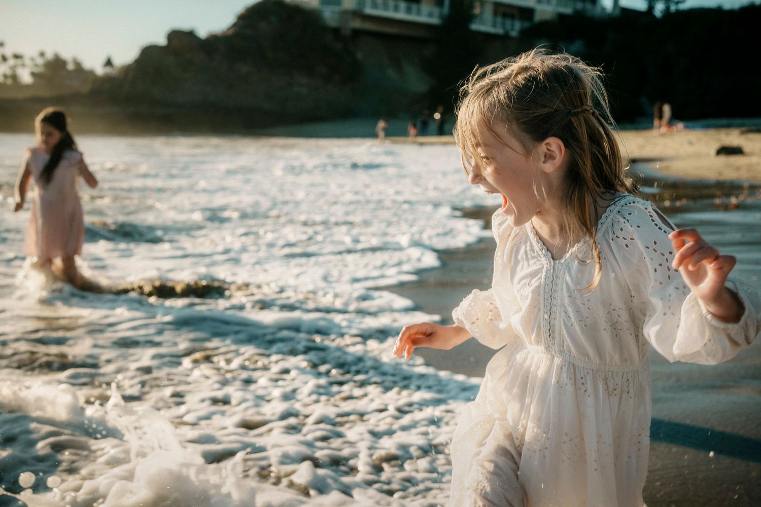 A young girl in a white dress plays in ocean waves on a sunny beach.