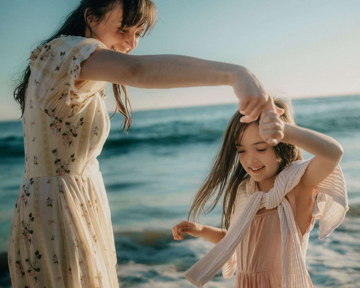 Mother twirling her smiling daughter on a beach with the ocean in the background.