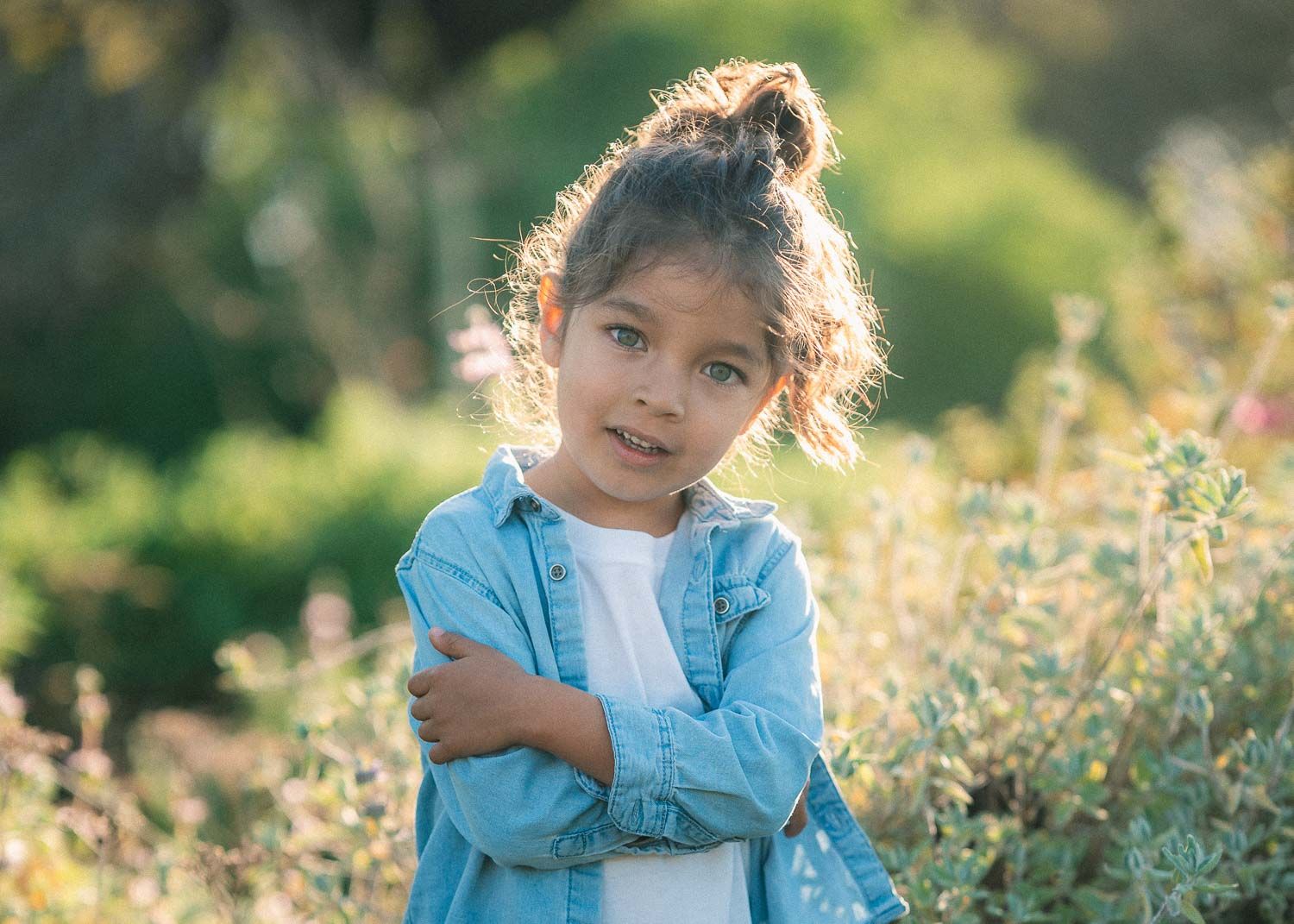 Young girl in denim shirt with arms crossed smiling in a sunny garden.