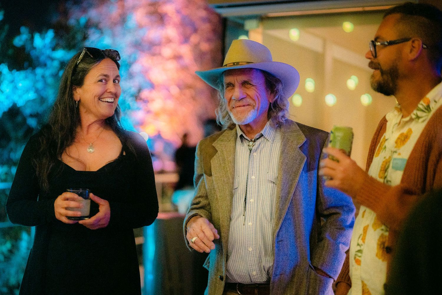 Three guests laughing and socializing at a vibrant outdoor evening party under colorful lights.