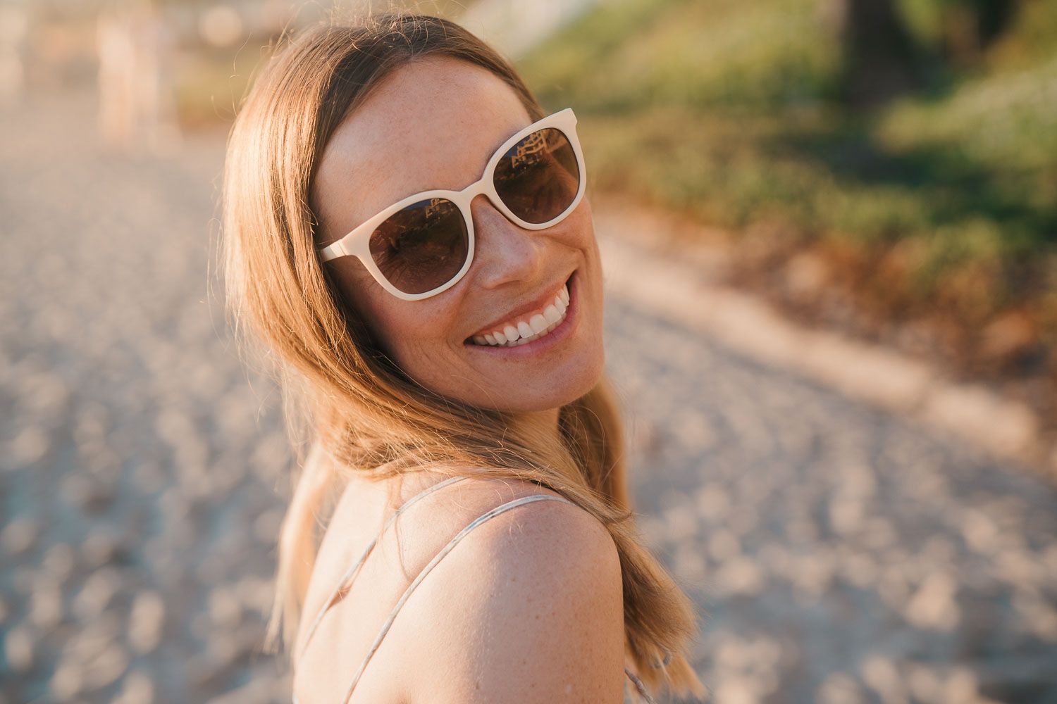 Smiling blonde woman wearing white sunglasses at a sunny beach location.