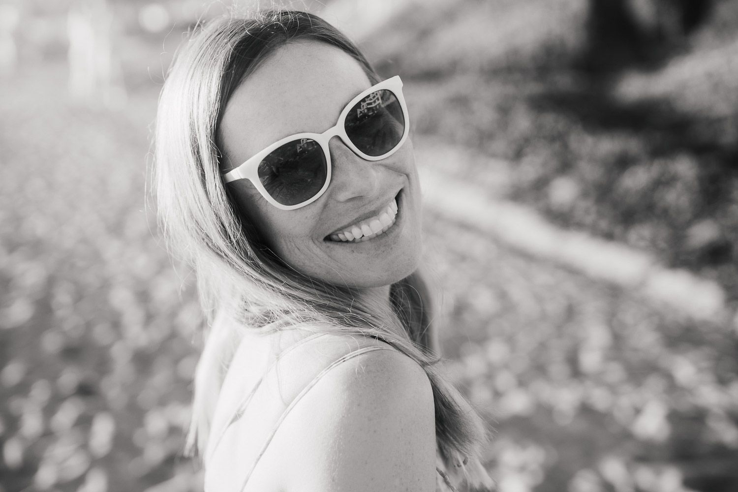 Black and white portrait of a smiling blonde woman wearing white sunglasses outdoors.