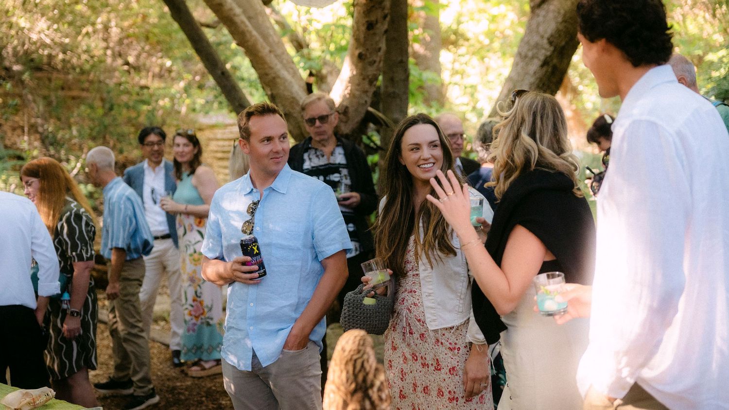 Guests laugh and socialize at a sunlit outdoor garden party surrounded by trees and greenery.