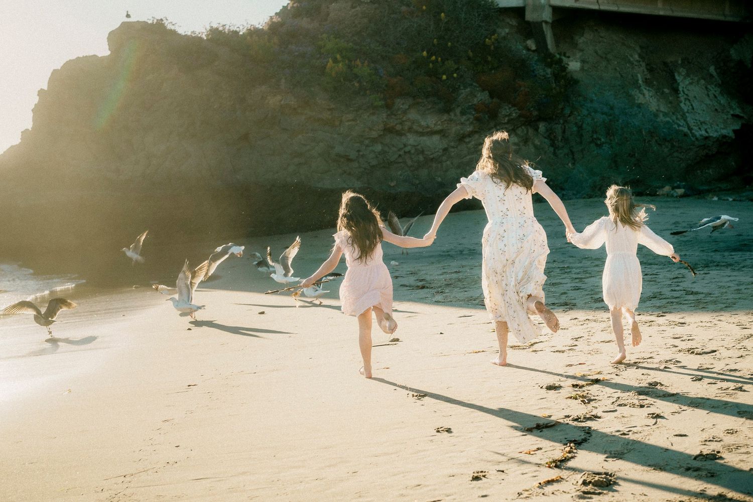 Woman and two girls running hand-in-hand on a sunlit beach with seagulls.