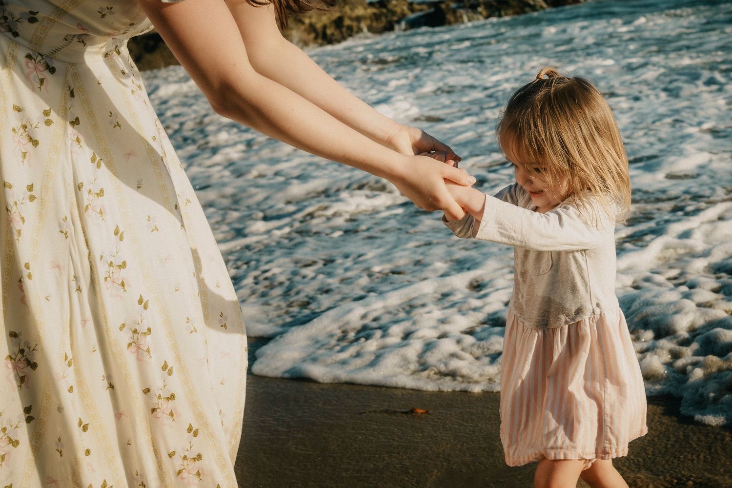 Young toddler girl in pink dress holding mothers hands while playing near ocean waves on a sandy beach.