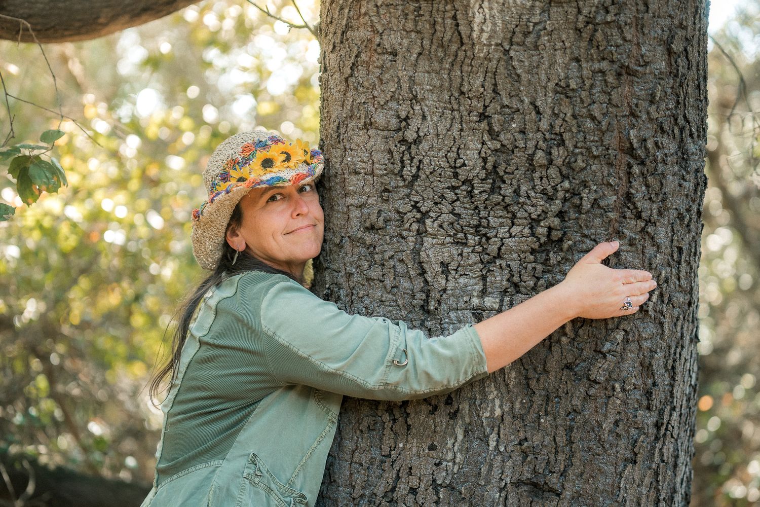 Woman in colorful sunflower hat hugging a large tree trunk in a sunlit forest setting.