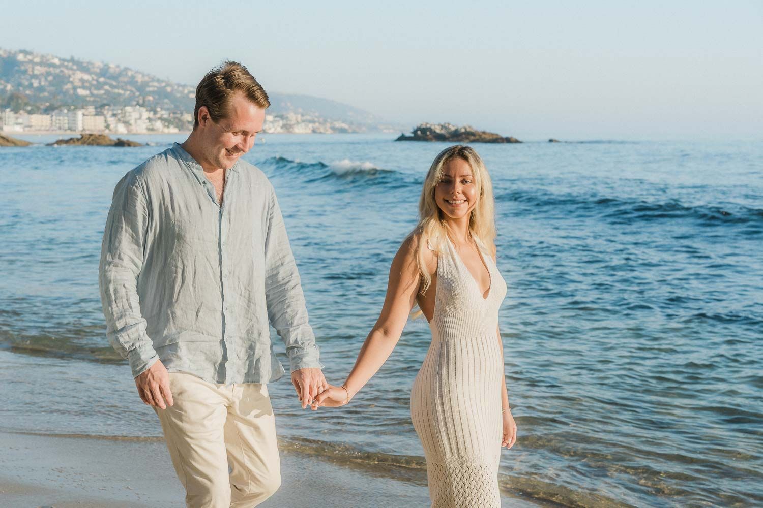 Happy couple walking hand in hand along sunny beach shoreline with blue ocean water in background.