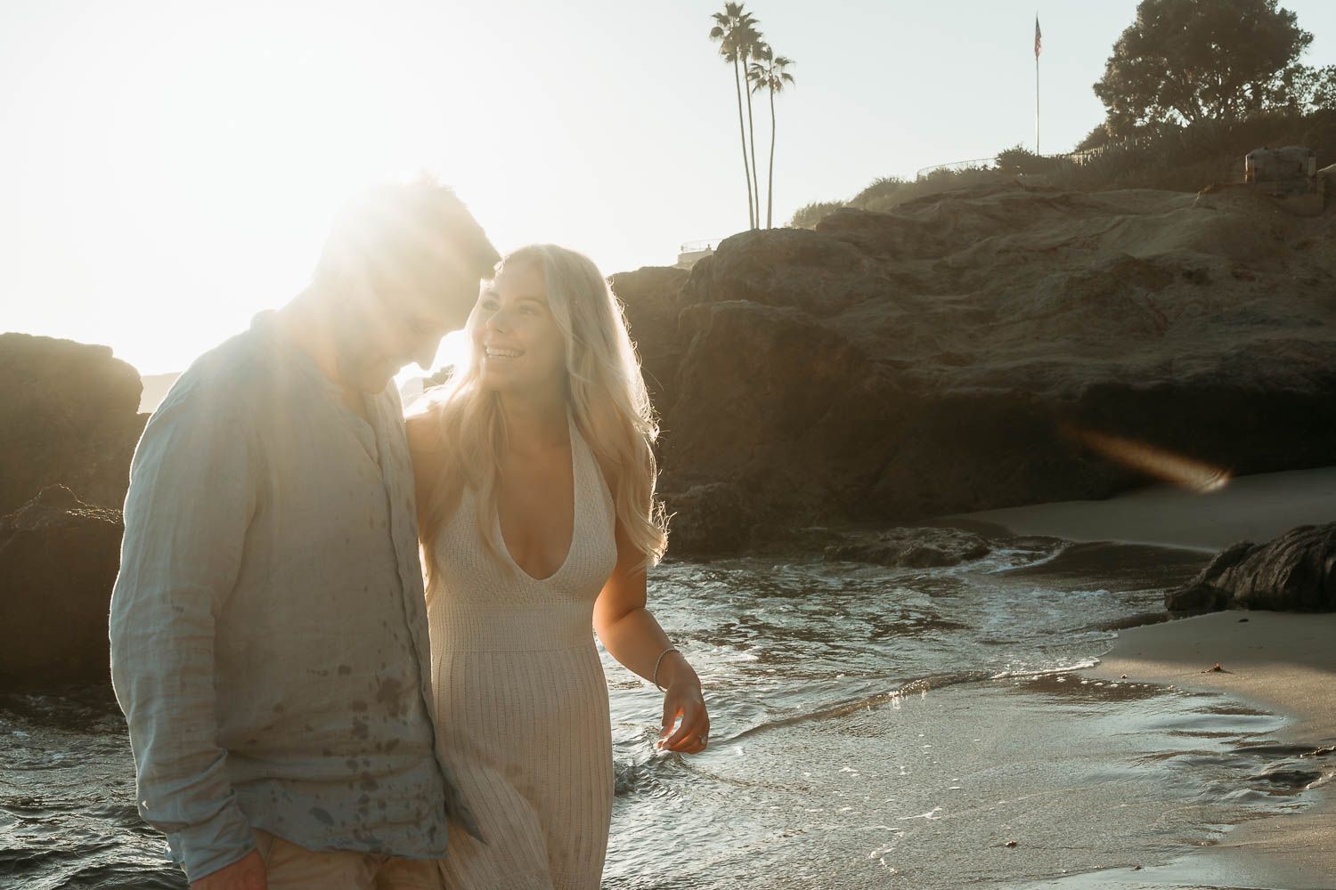 Romantic couple walking closely together on a sunlit beach, woman in white dress near rocky coastal cliffs.