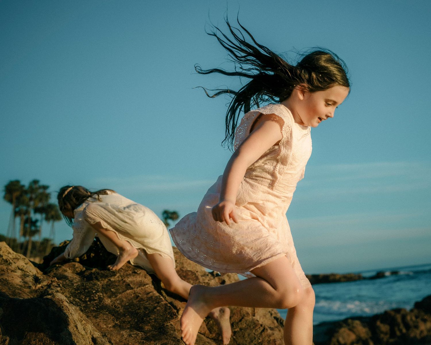 Young girl in pink dress climbing rocks on a sunny beach with wind in her hair.