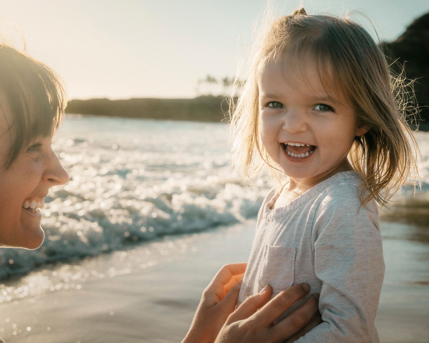 A joyful toddler girl laughs on a sunny beach while being held by a smiling adult.