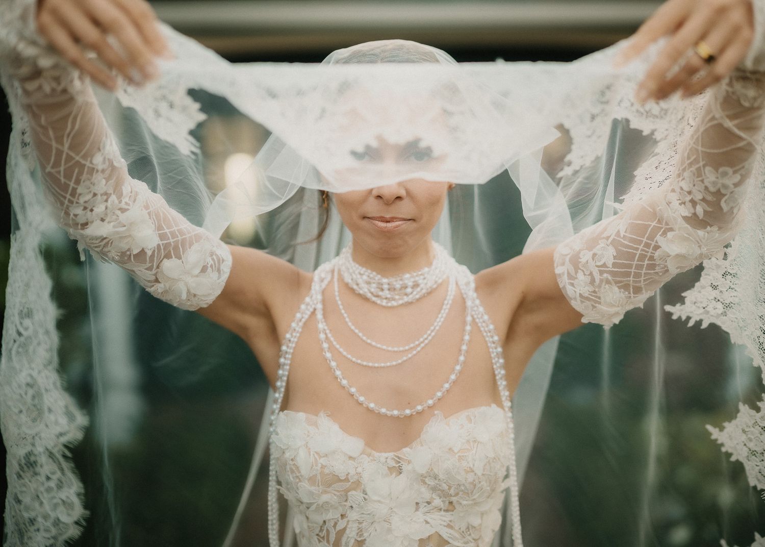 A bride lifts her lace veil overhead wearing pearl necklaces and an elegant strapless gown.