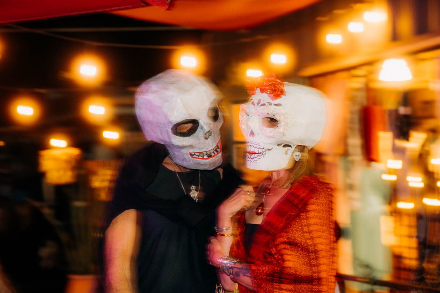 Two people wearing skull masks celebrate Day of the Dead at a festive Halloween party with warm glowing lights.