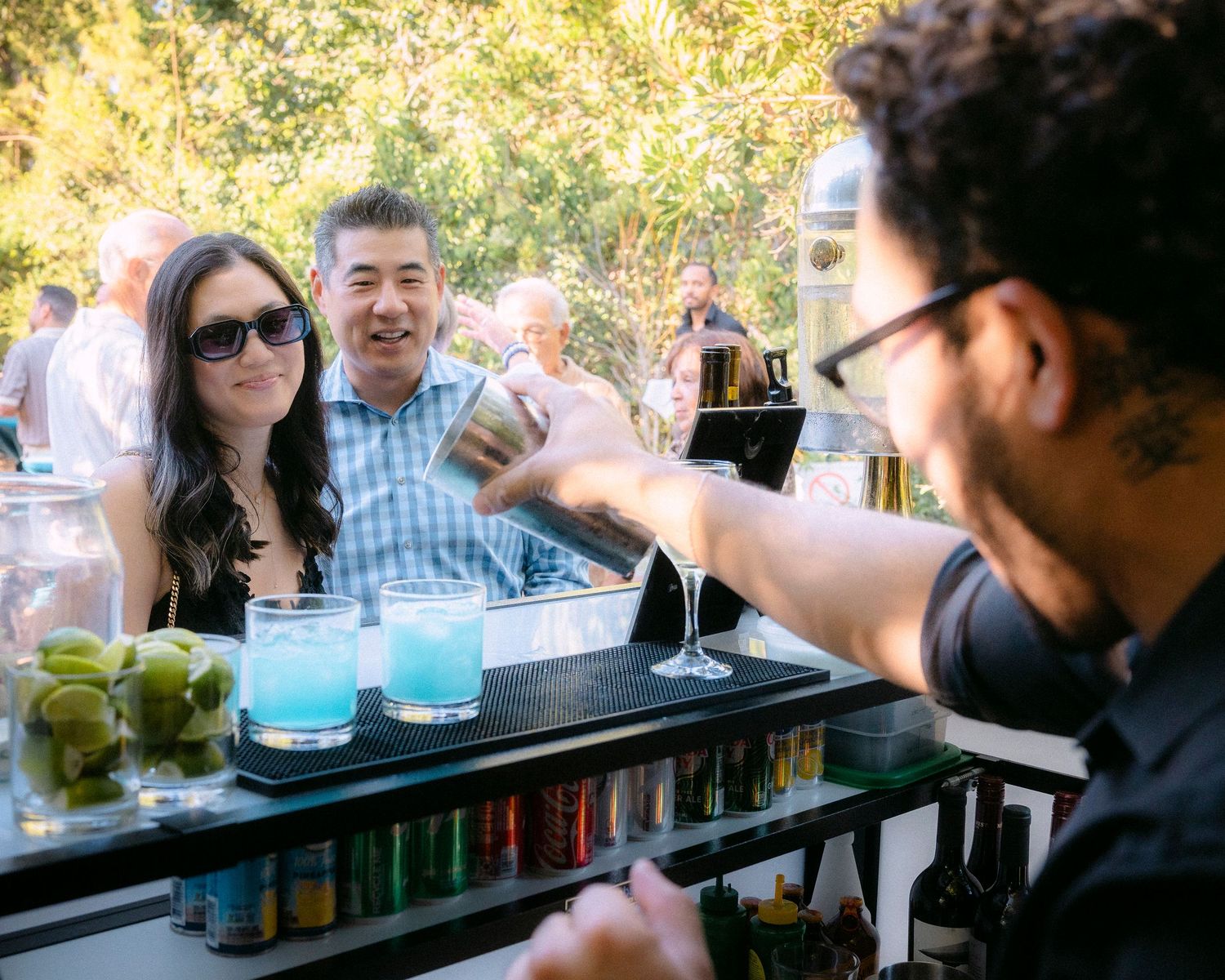 Bartender shakes cocktails for smiling couple at an elegant outdoor bar with blue drinks and fresh limes.