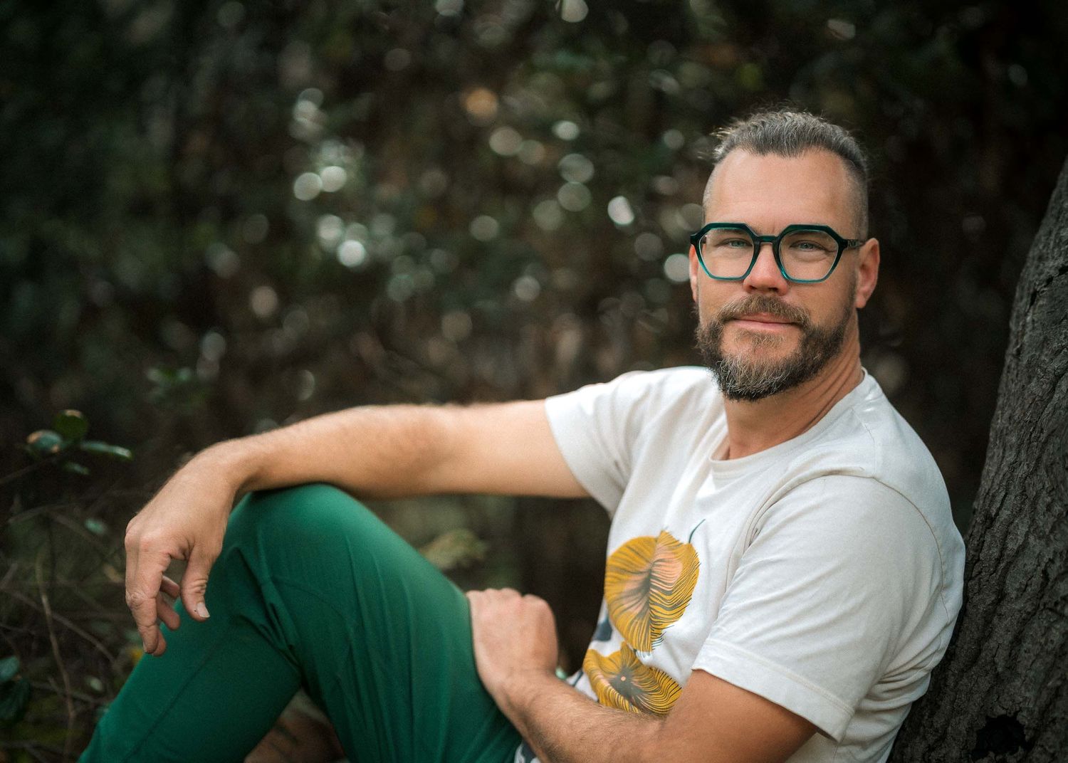 Bearded man in green pants and white t-shirt relaxes against a tree in a lush outdoor setting.