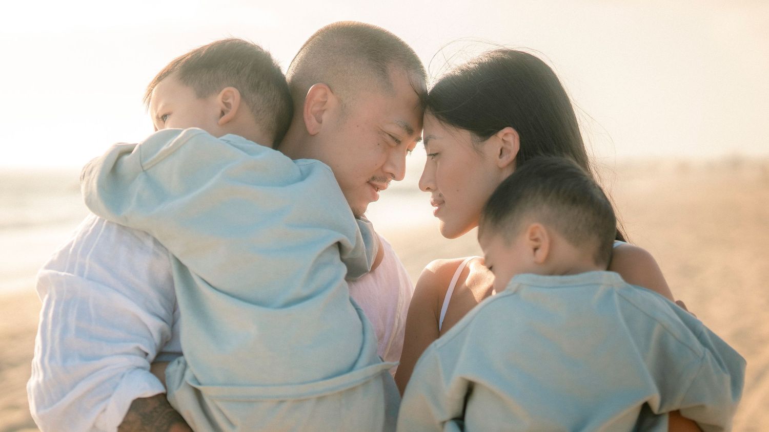 A couple sharing a tender moment on a beach at sunset while each holding a baby in their arms.