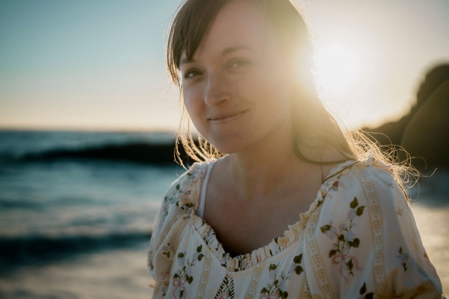 Woman with dark hair smiling softly at sunset on a beach with golden backlight.