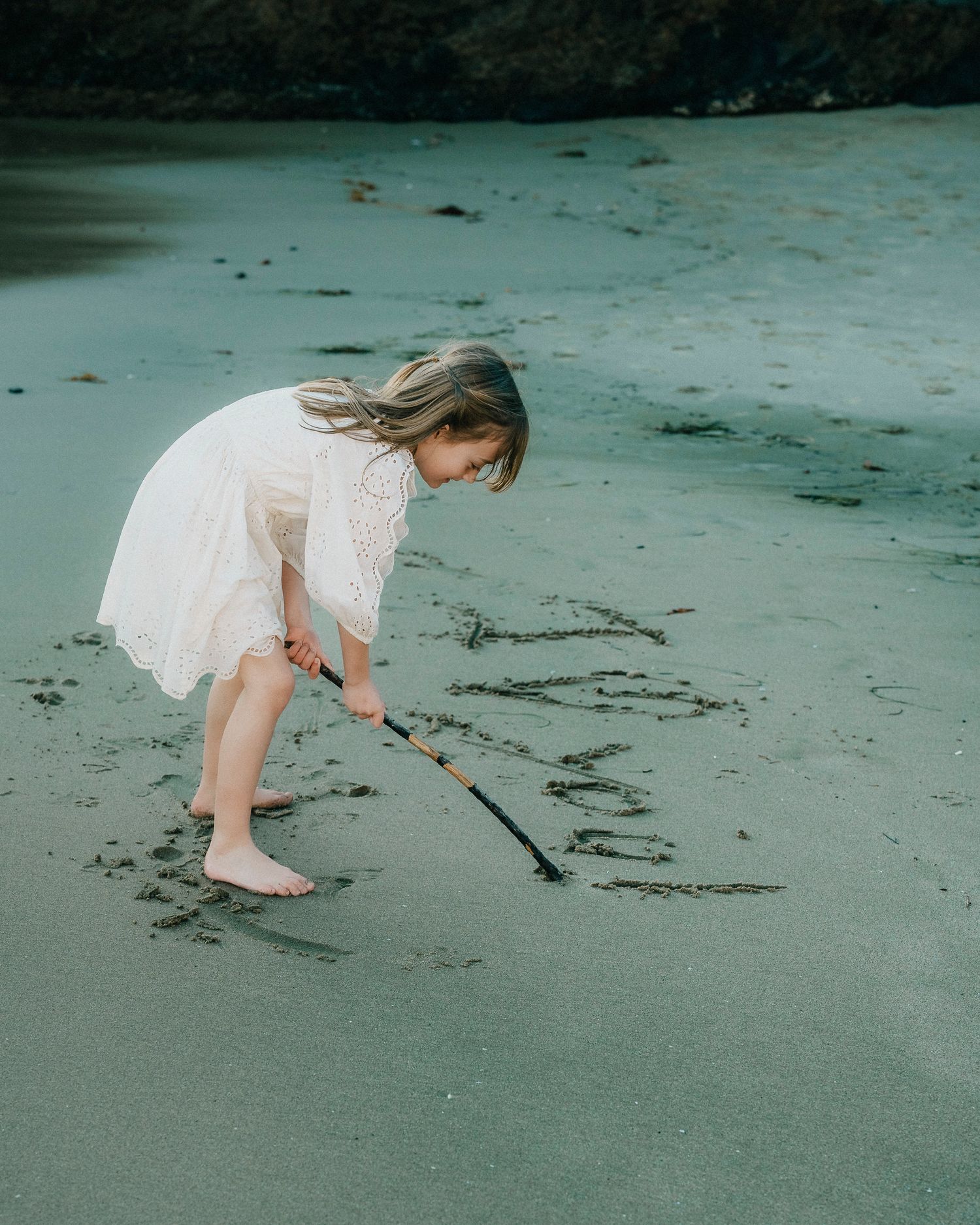 Young girl in white dress writing in wet sand on a beach.