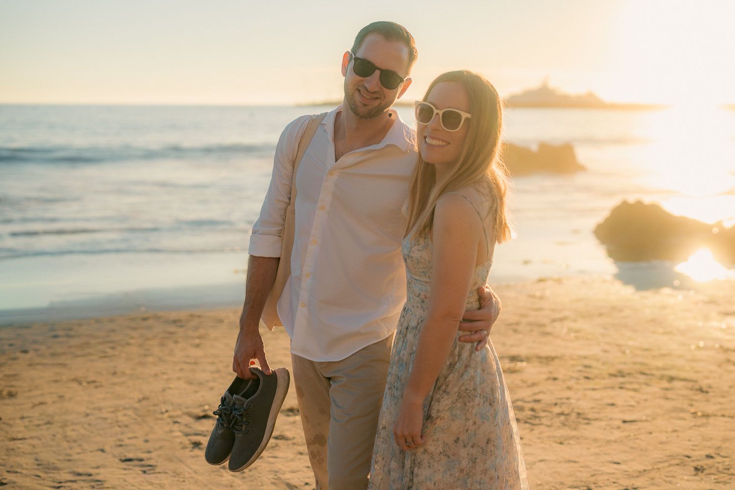Smiling couple in sunglasses walking barefoot on sandy beach at sunset, man carrying shoes in hand.