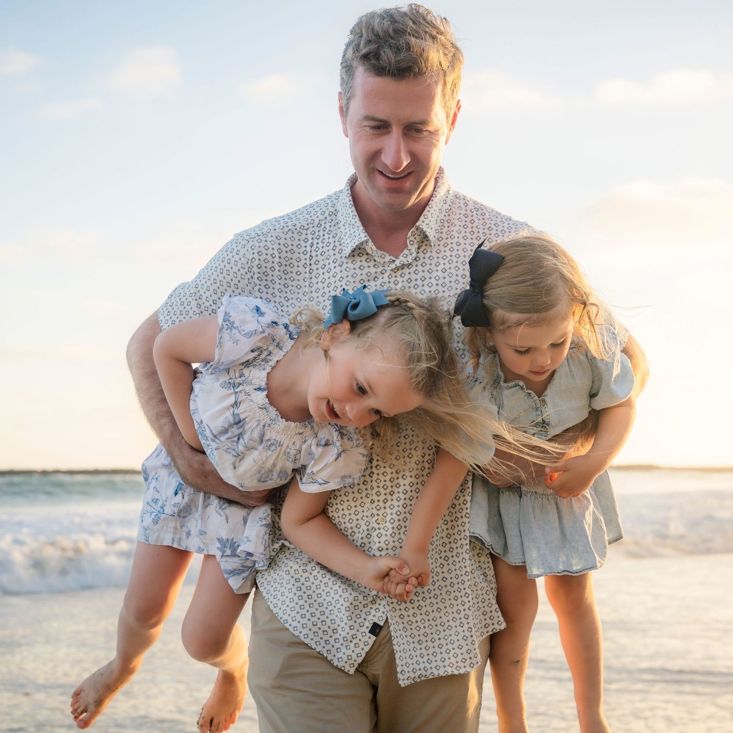 Father laughing while holding two young daughters on a sunny beach at golden hour.