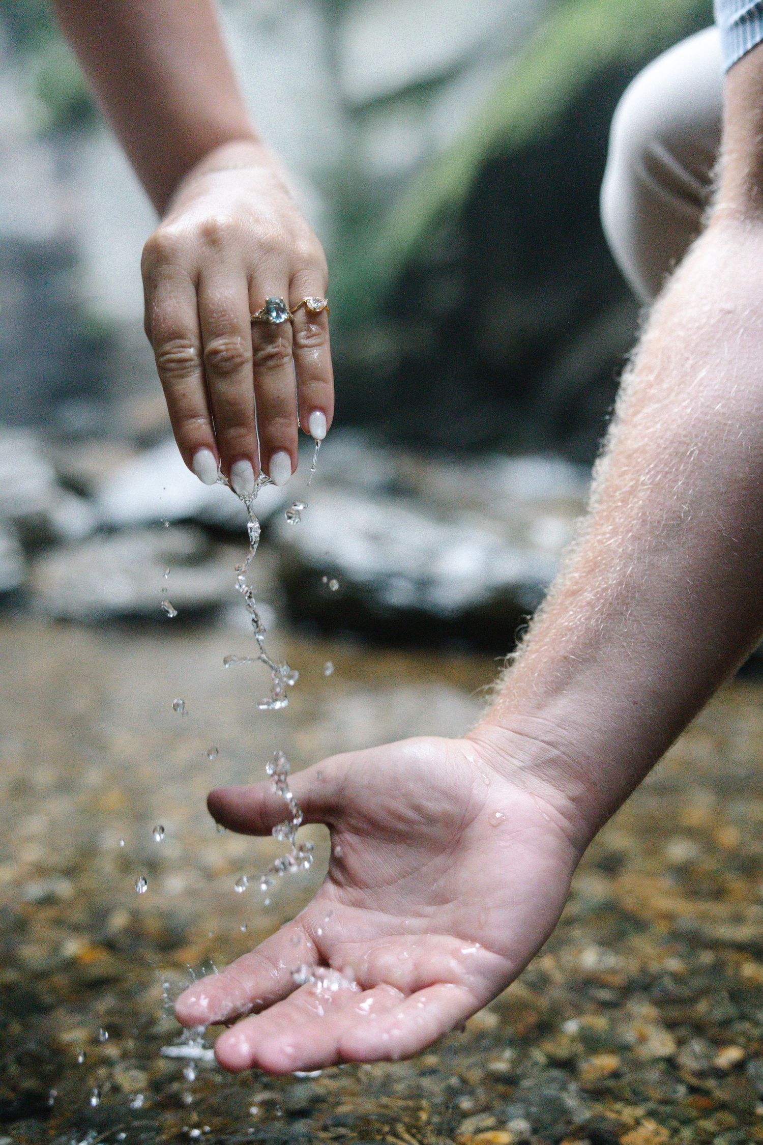 Water droplets falling from a woman's ringed hand into a man's cupped palm near a rocky stream.