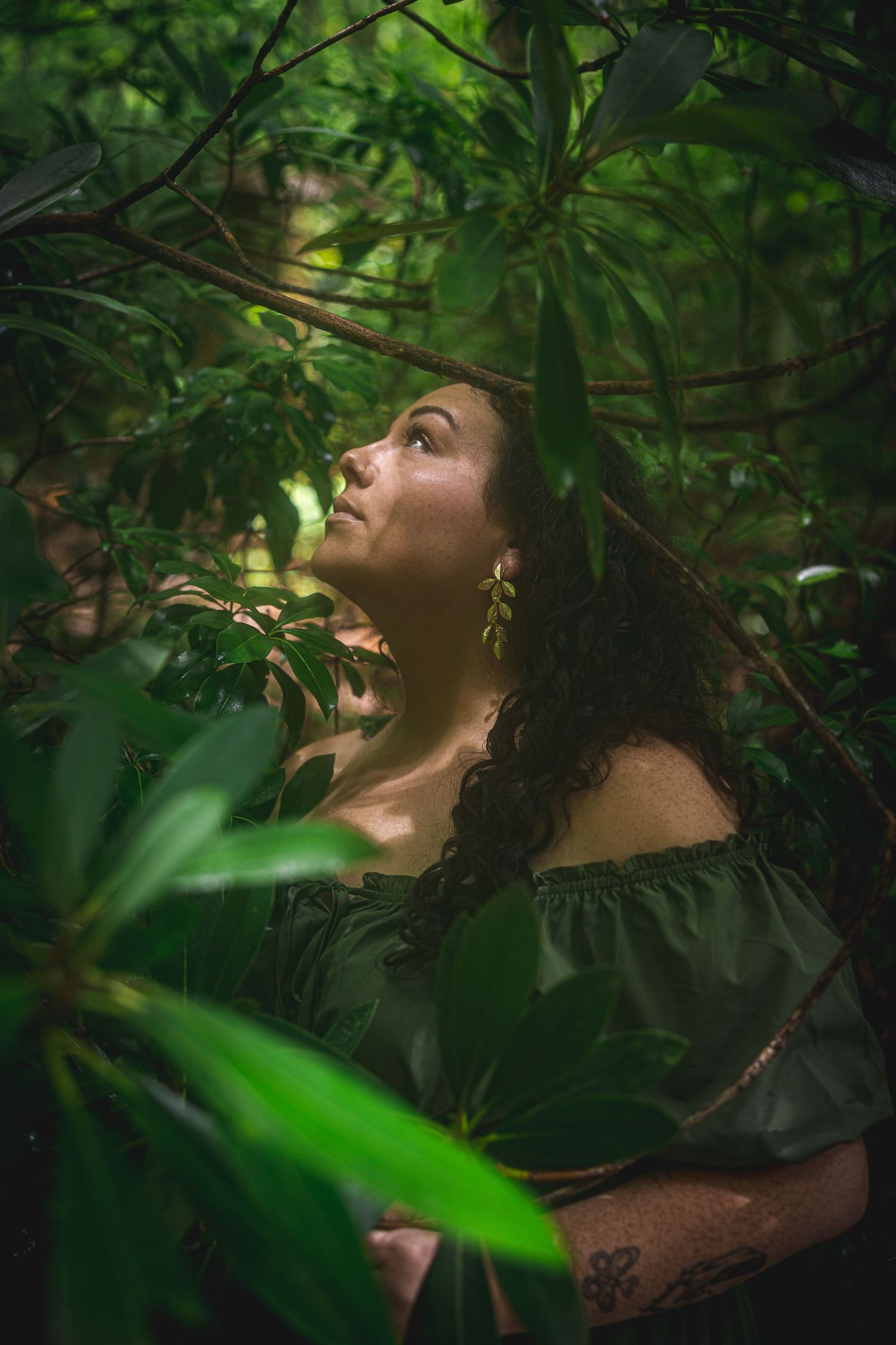 Curly-haired woman in green off-shoulder dress gazing upward through lush forest foliage.