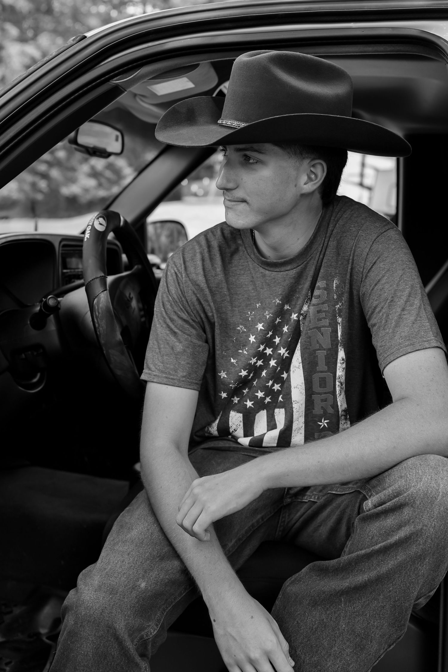 Young man in cowboy hat and senior t-shirt sits in truck door in black and white portrait photo.