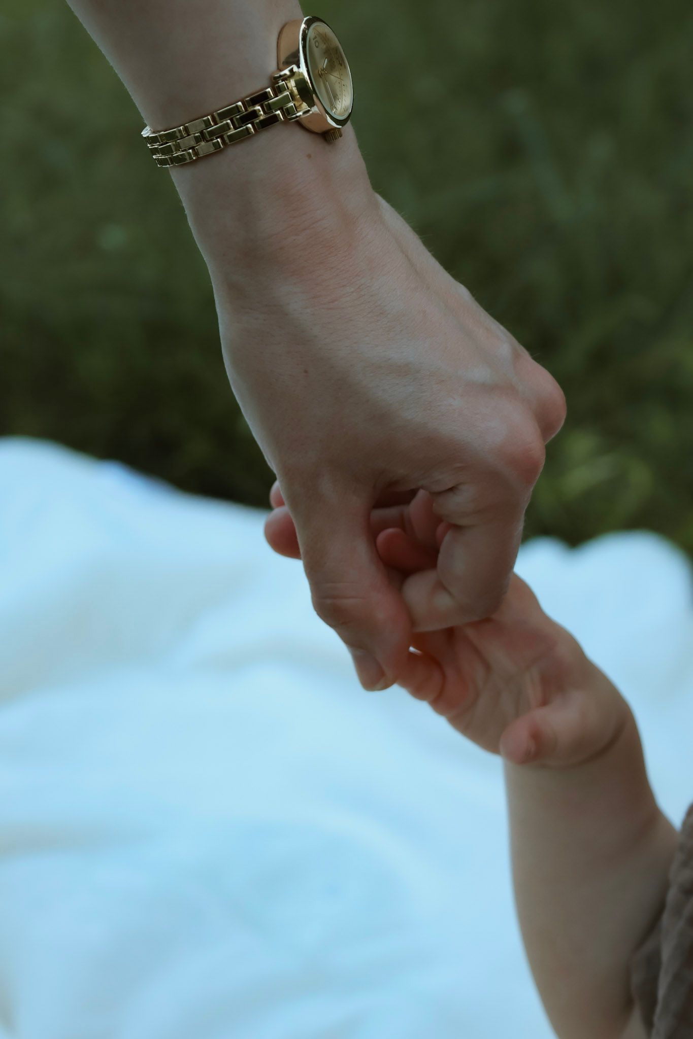 A mother wearing a gold watch gently holds her baby's tiny hand on a white blanket outdoors.