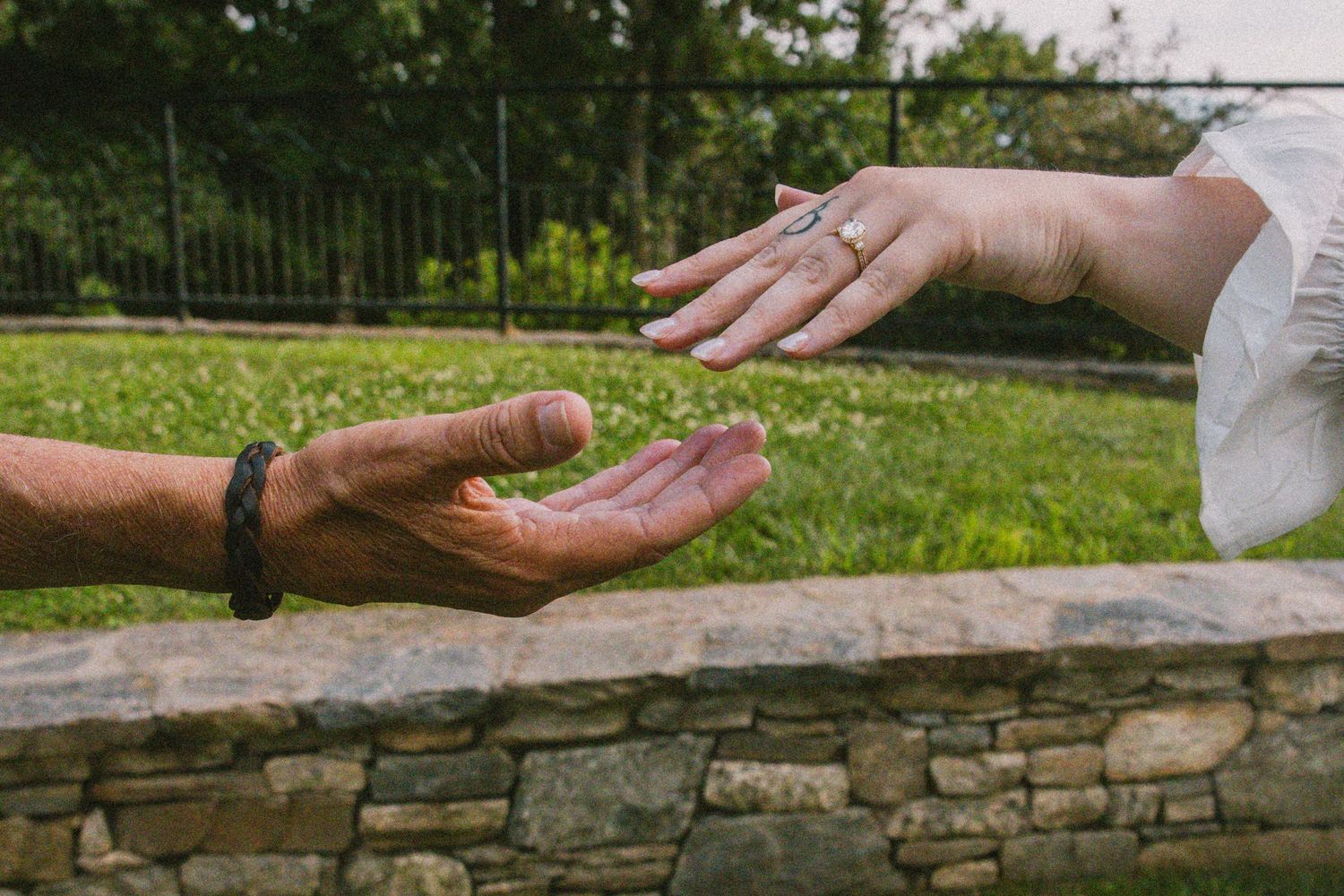 Two hands reaching toward each other near a stone wall, one wearing a diamond ring and the other a black bracelet.