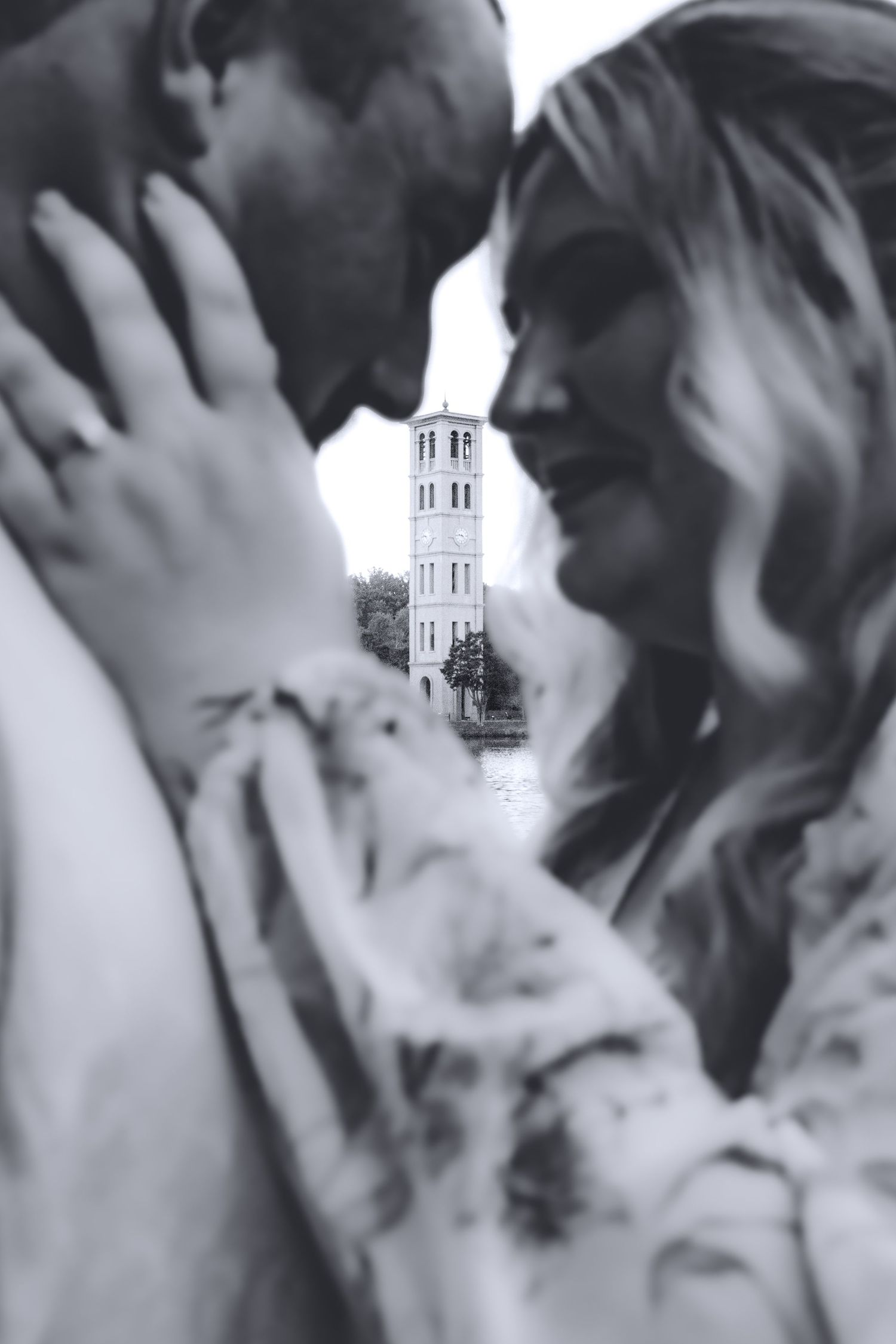 Black and white photo of a couple leaning in close with a historic tower reflected in water visible between them.