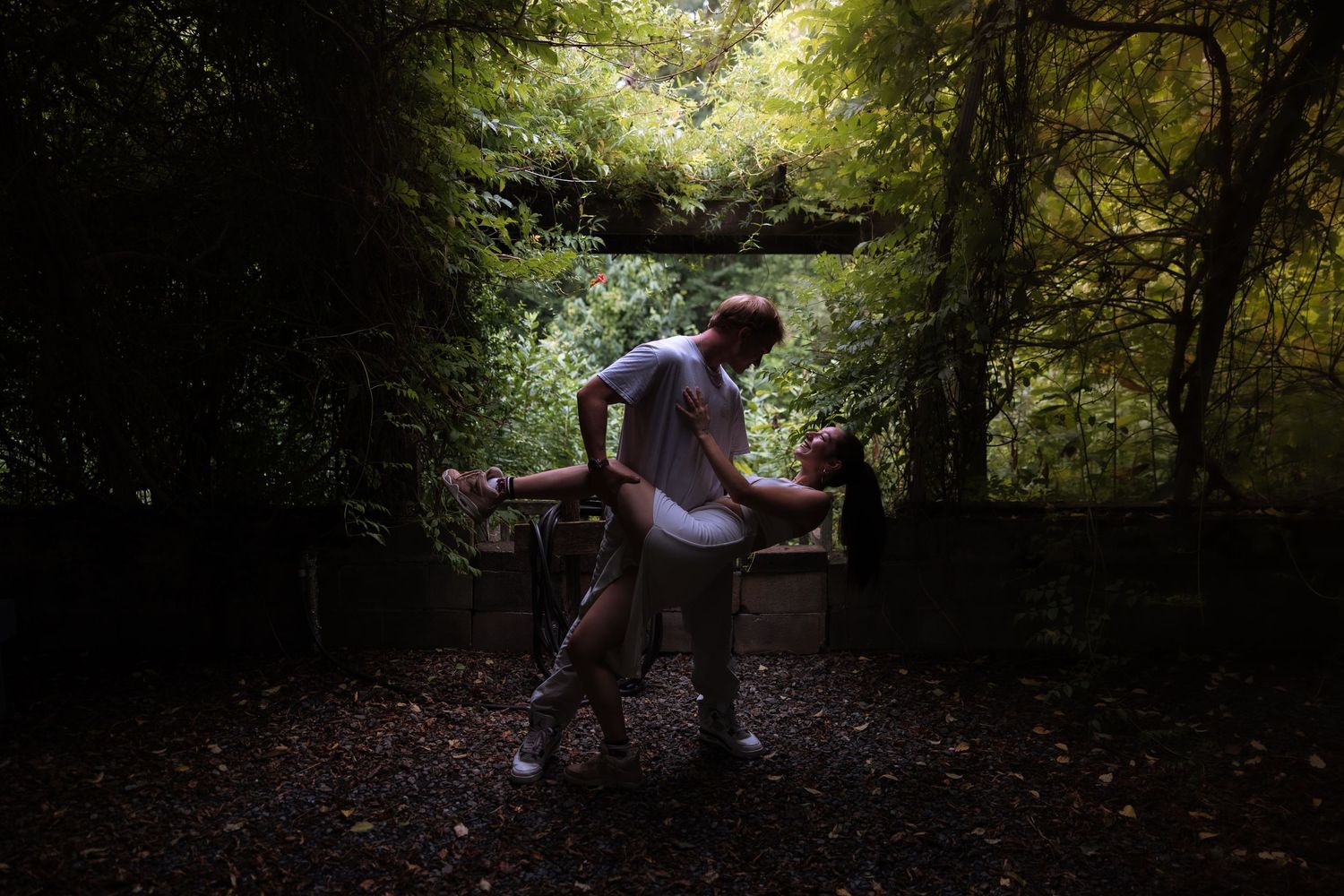 Couple dancing playfully on a dark forest path surrounded by lush green trees.