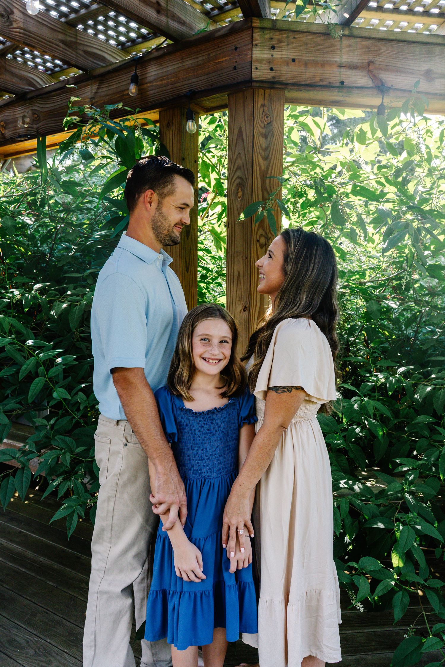Happy family of three smiling together under a wooden pergola surrounded by tropical greenery.