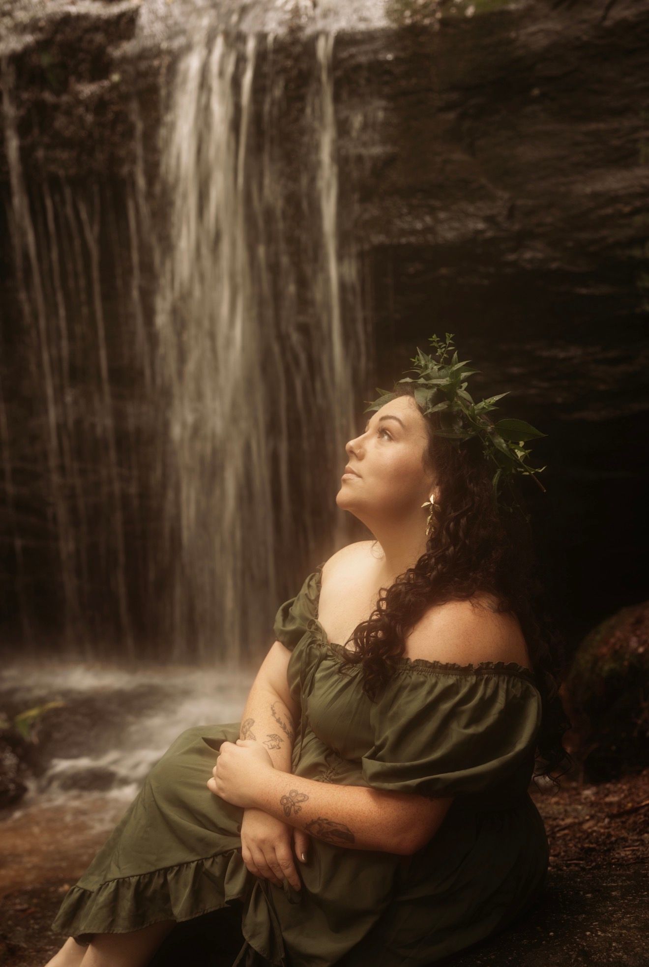 Woman in green off-shoulder dress with floral crown sitting peacefully before a waterfall.