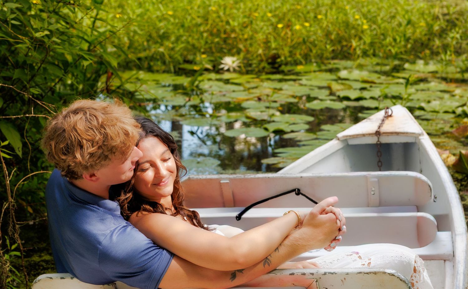 Romantic couple embracing in a white rowboat surrounded by lily pads on a serene pond.