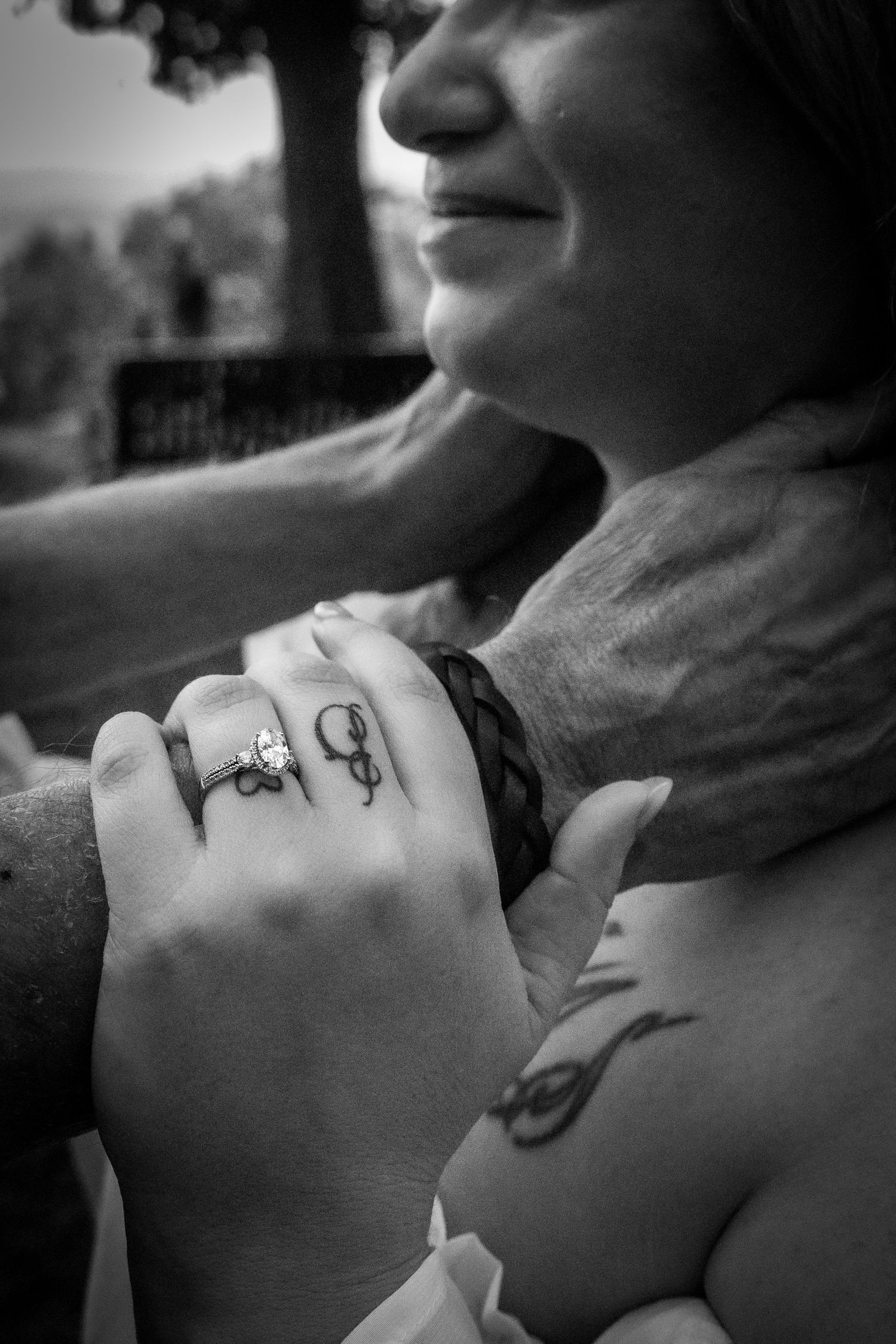 Black and white photo of a smiling woman with tattoos and rings, embraced from behind by a man.