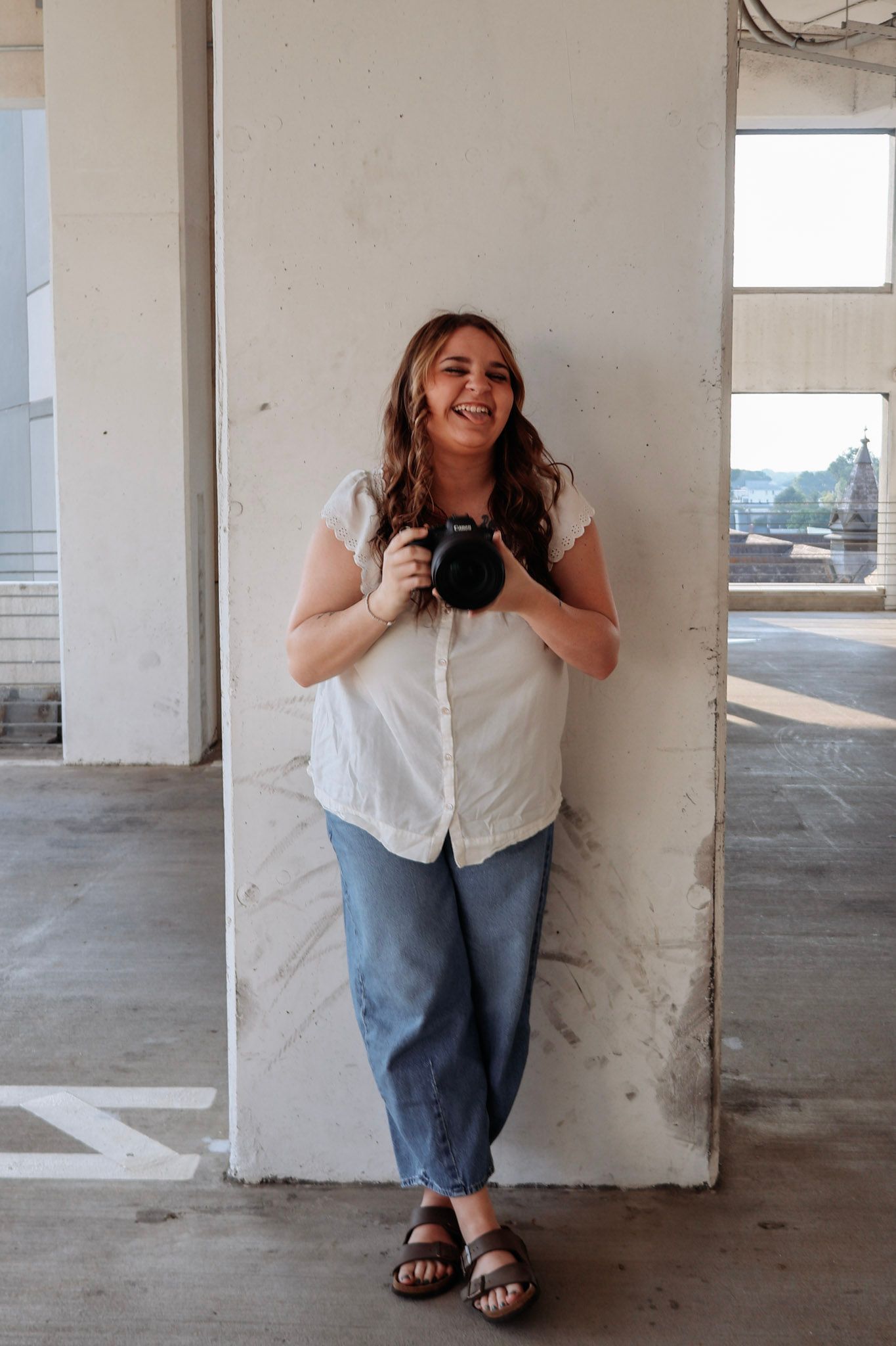 A smiling woman leans against a concrete pillar holding a DSLR camera in a parking structure.
