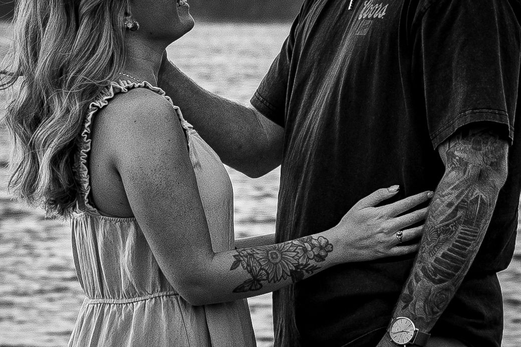 Close-up of a tattooed couple embracing near a lake in black and white.