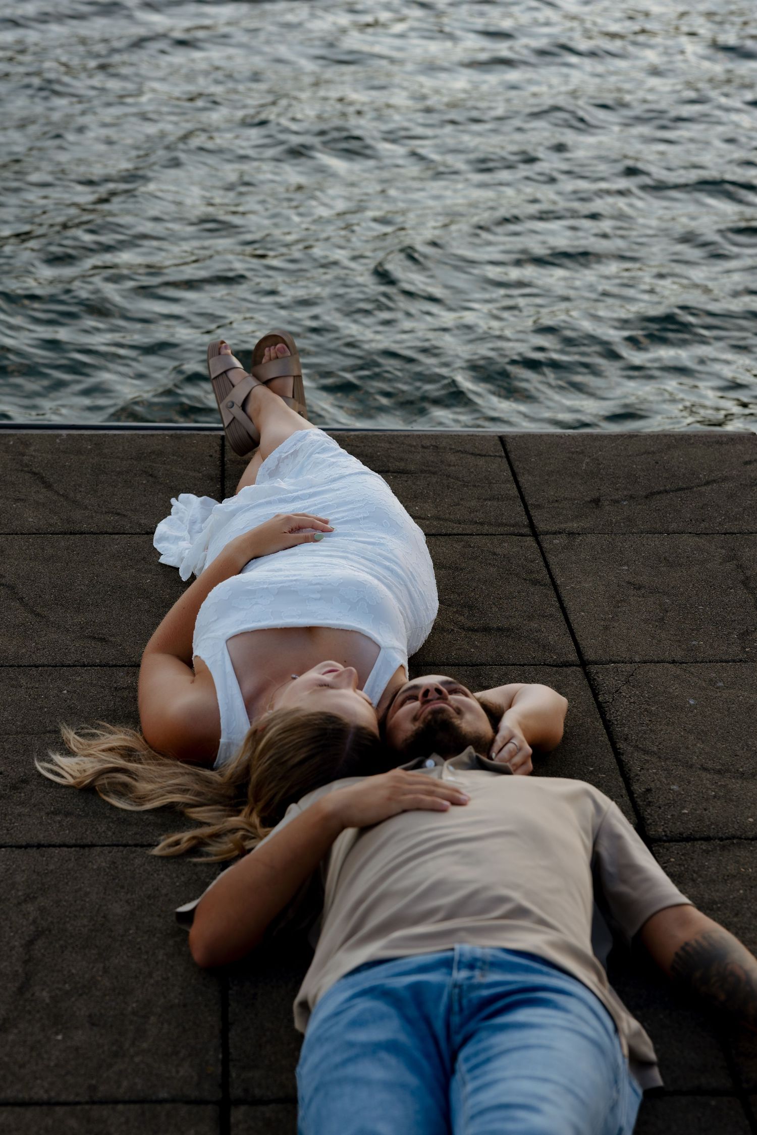 Couple lying on a waterfront pier, woman in white dress and man in jeans, sharing a romantic moment by the water.