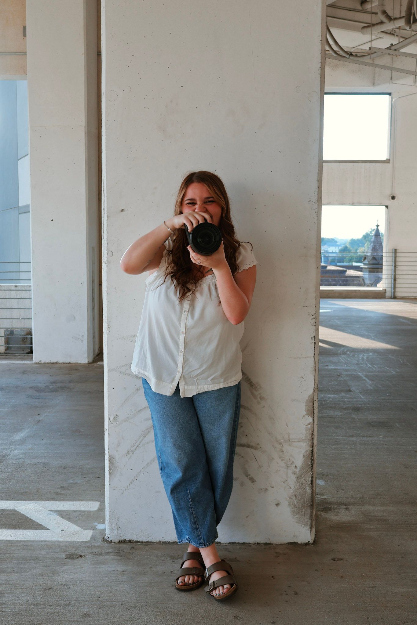 Young woman in white shirt and jeans holding a camera up to her face in a parking garage.