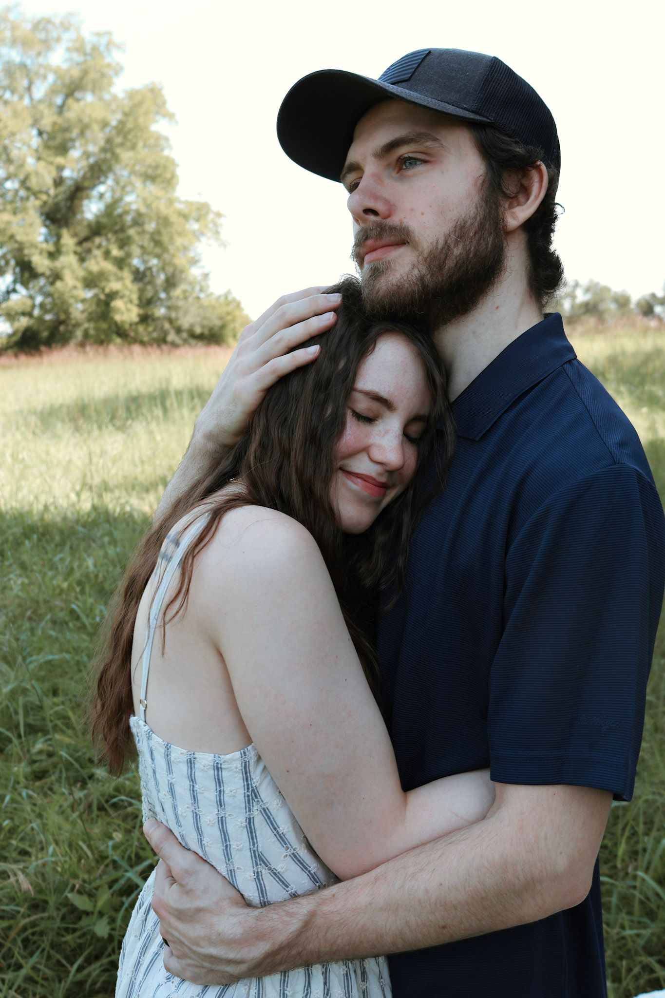 A couple embraces tenderly in a green field, the woman resting her head on the man's chest.