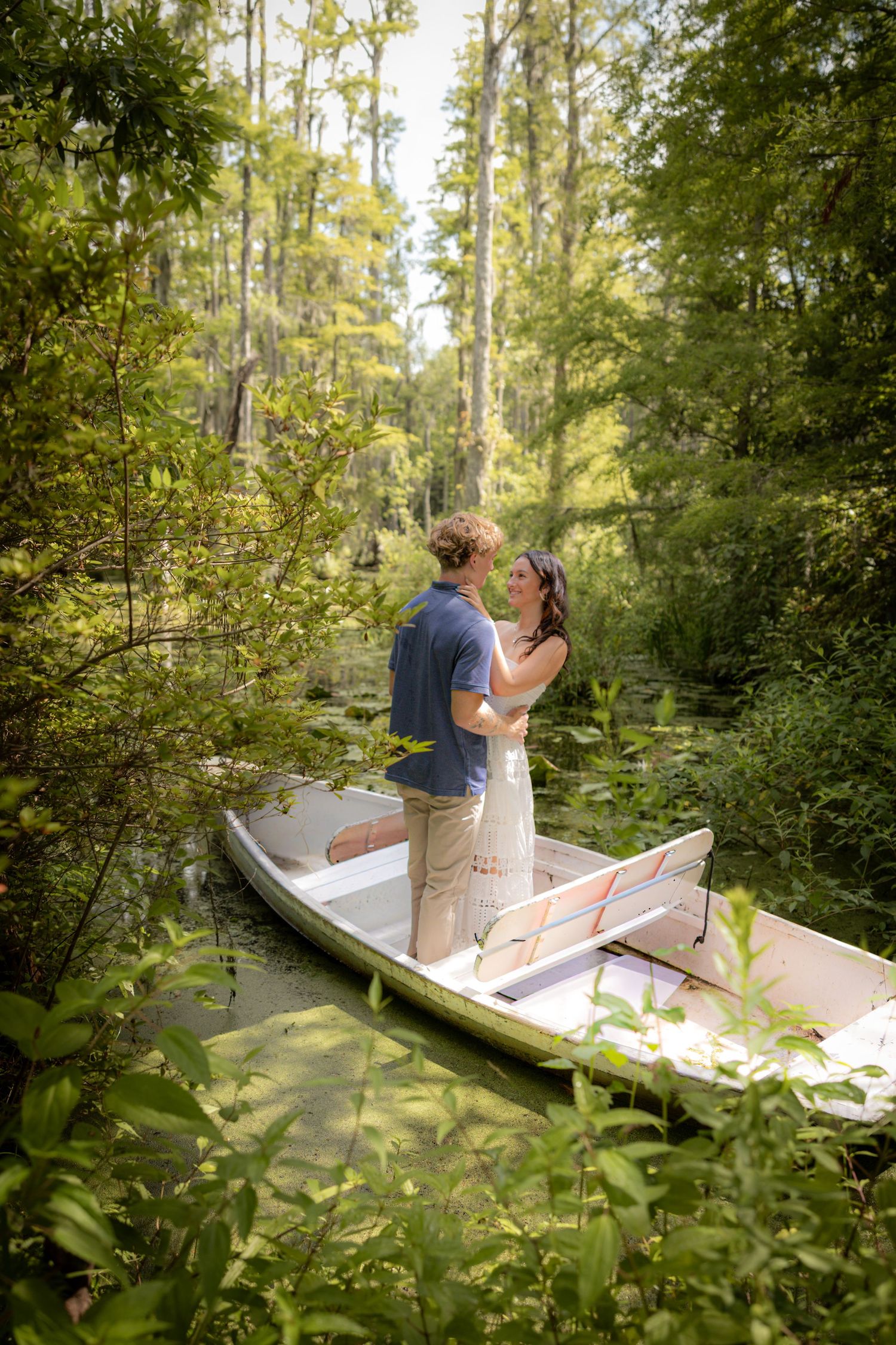 Couple standing and embracing in a small boat surrounded by lush green forest and calm water.