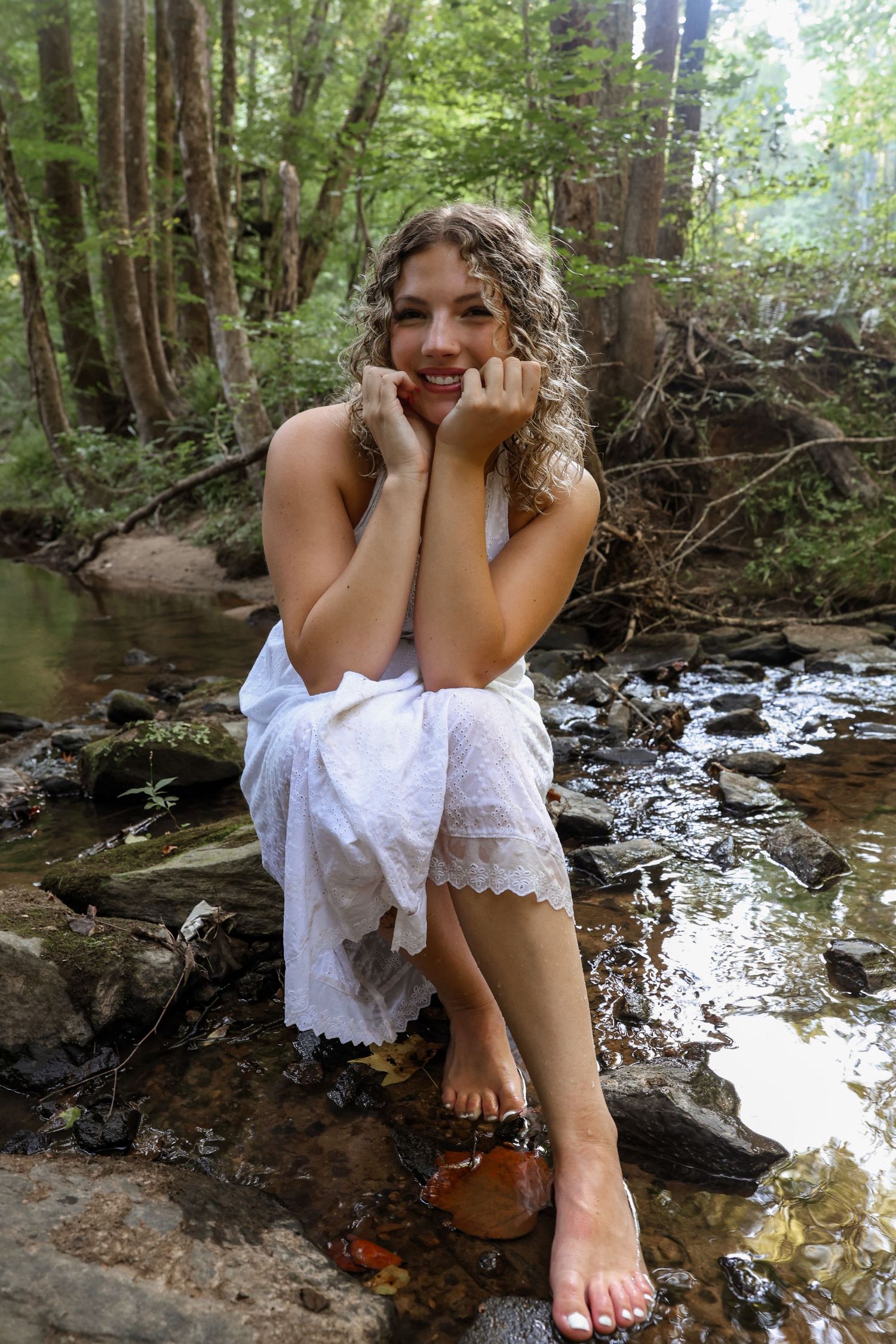 Smiling young woman with curly hair sitting barefoot on rocks beside a forest stream.