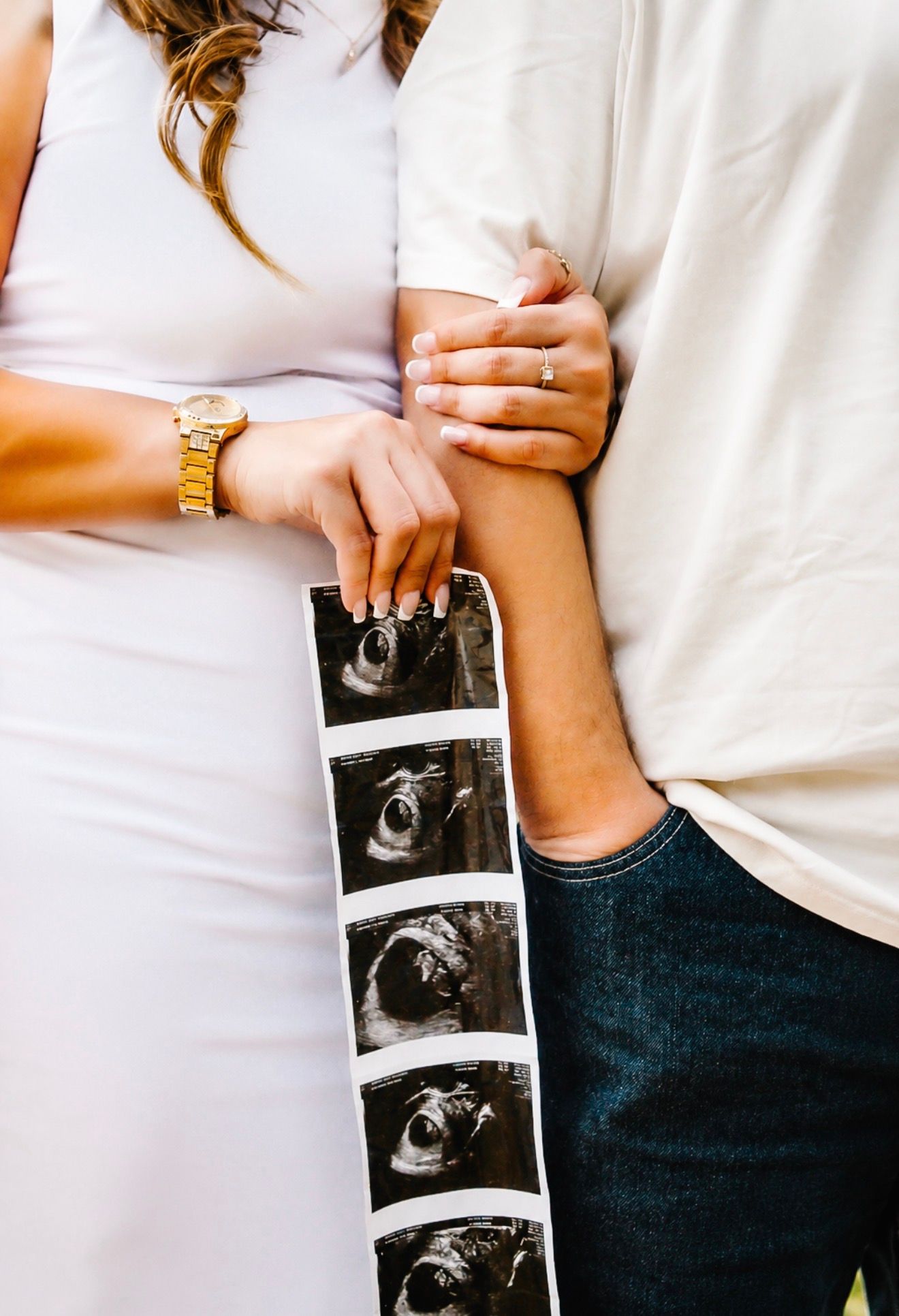 Expecting couple holding ultrasound photos together while standing close in coordinating white outfits.