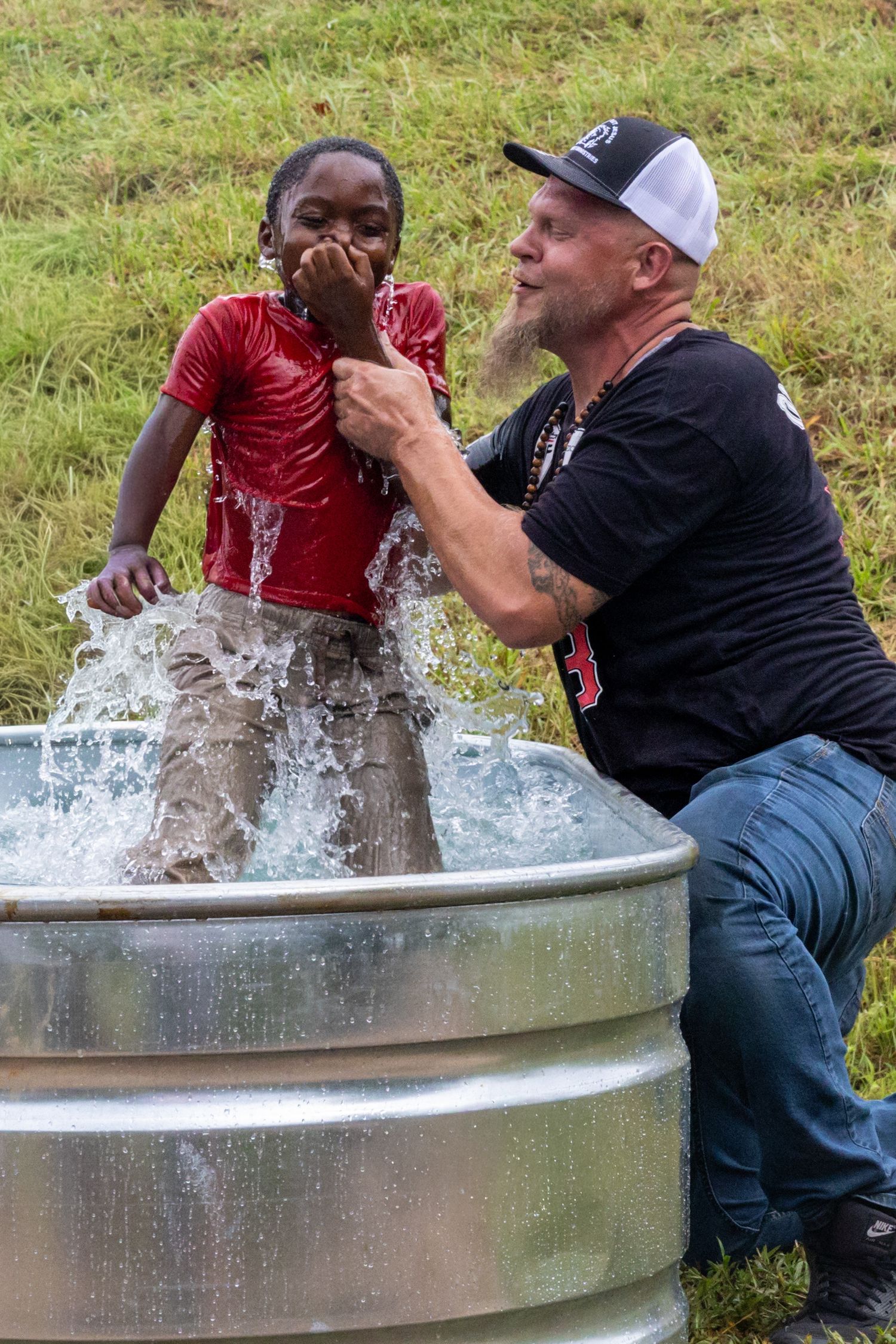 Man baptizing a young child in a metal water trough outdoors on a grassy field.