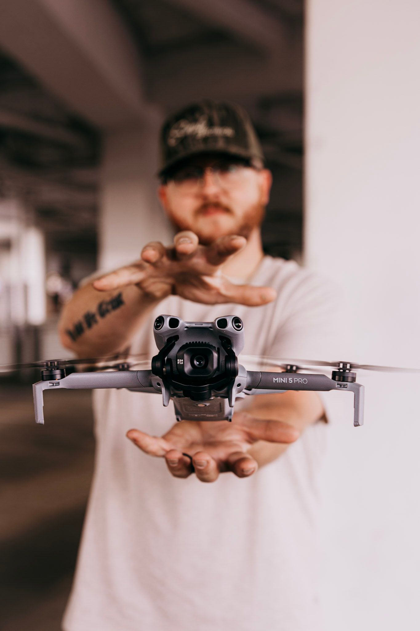 A tattooed man in a cap holds a DJI Mini 3 Pro drone toward the camera indoors.