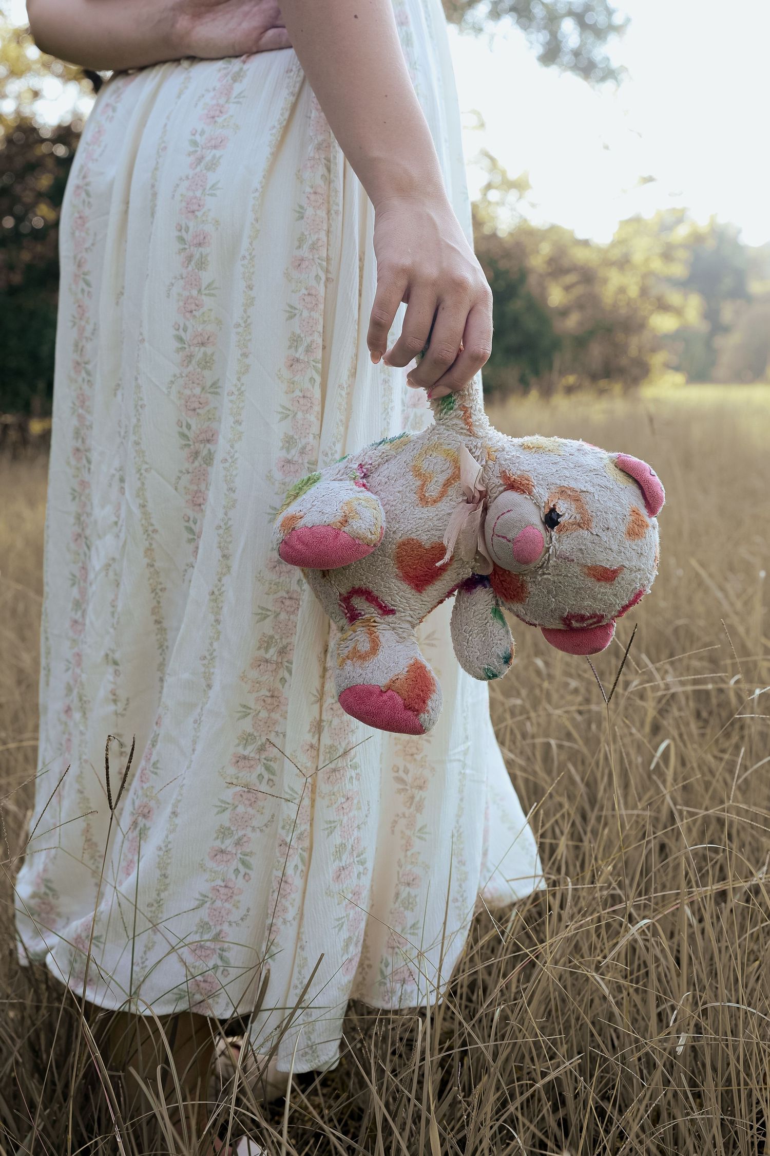 A woman in a floral dress holds a worn teddy bear in a golden field.