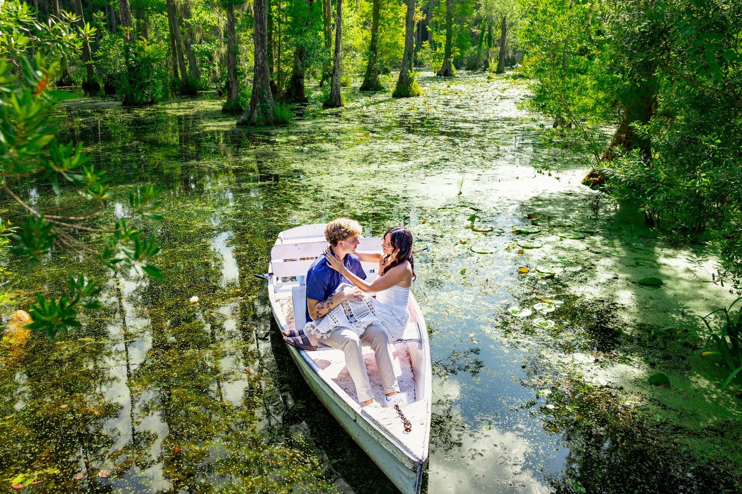 Couple sharing a romantic moment in a white rowboat on a serene, lush green swamp surrounded by cypress trees.