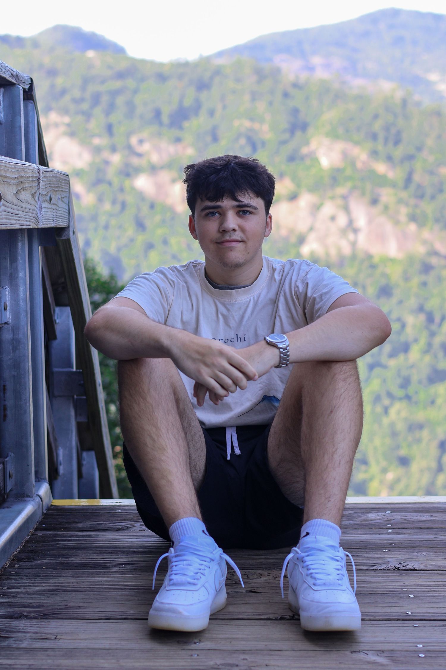 Young man sitting on wooden deck with arms crossed, wearing white sneakers and casual outfit, mountain forest backdrop.