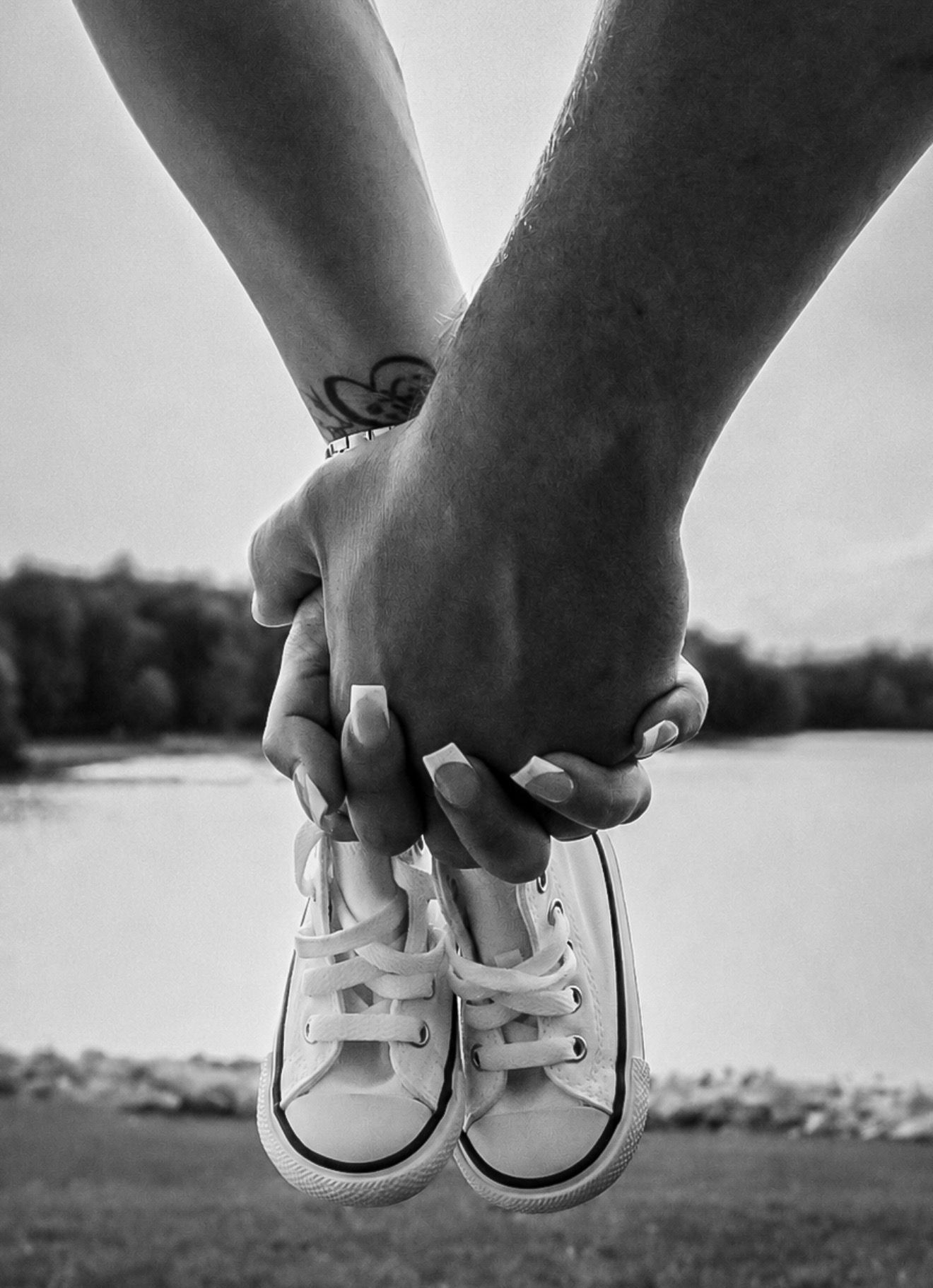 Black and white photo of couple holding hands with tiny baby Converse shoes near a lake.