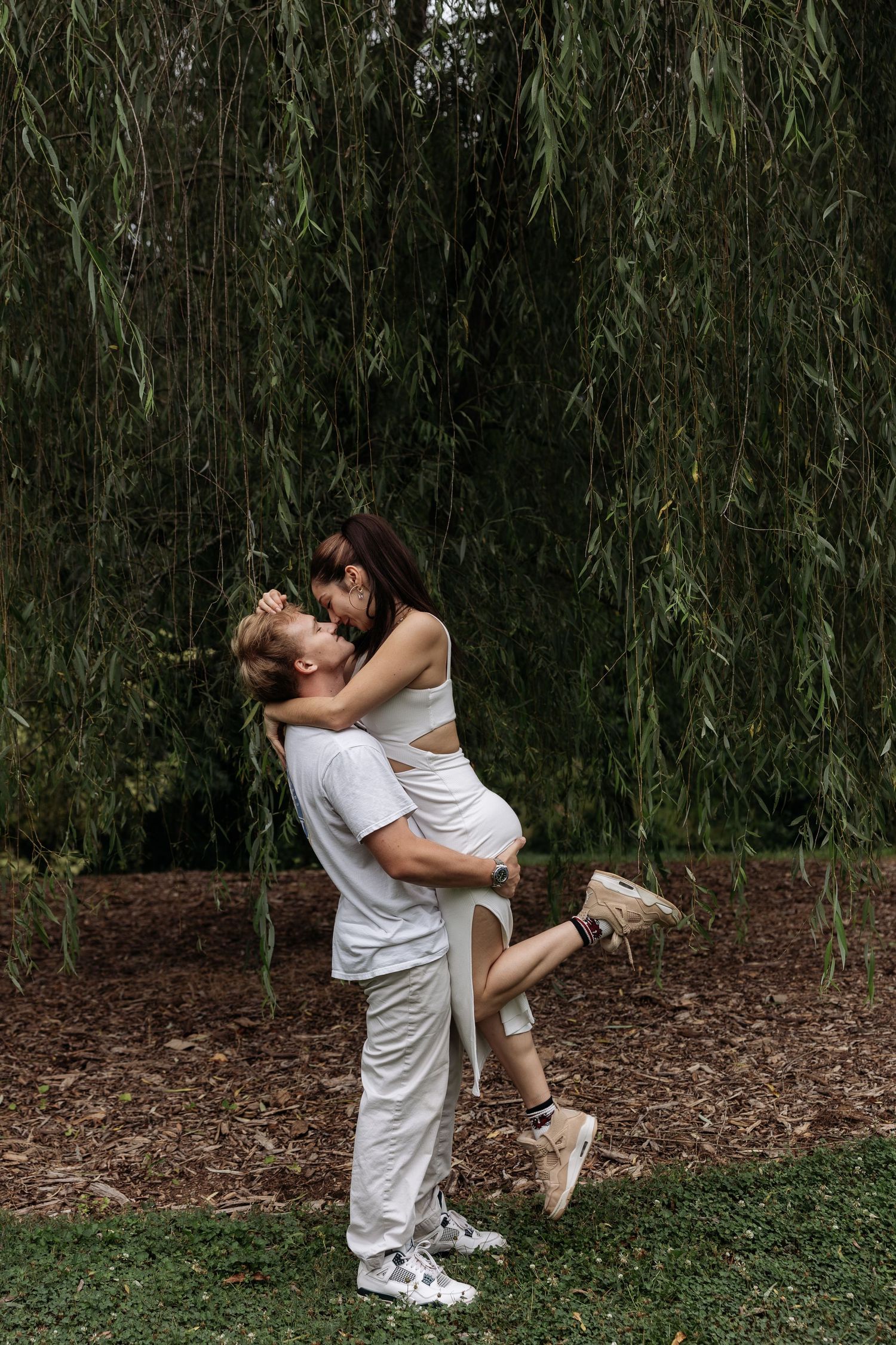 Man lifting and embracing pregnant woman in white dress under a weeping willow tree outdoors.
