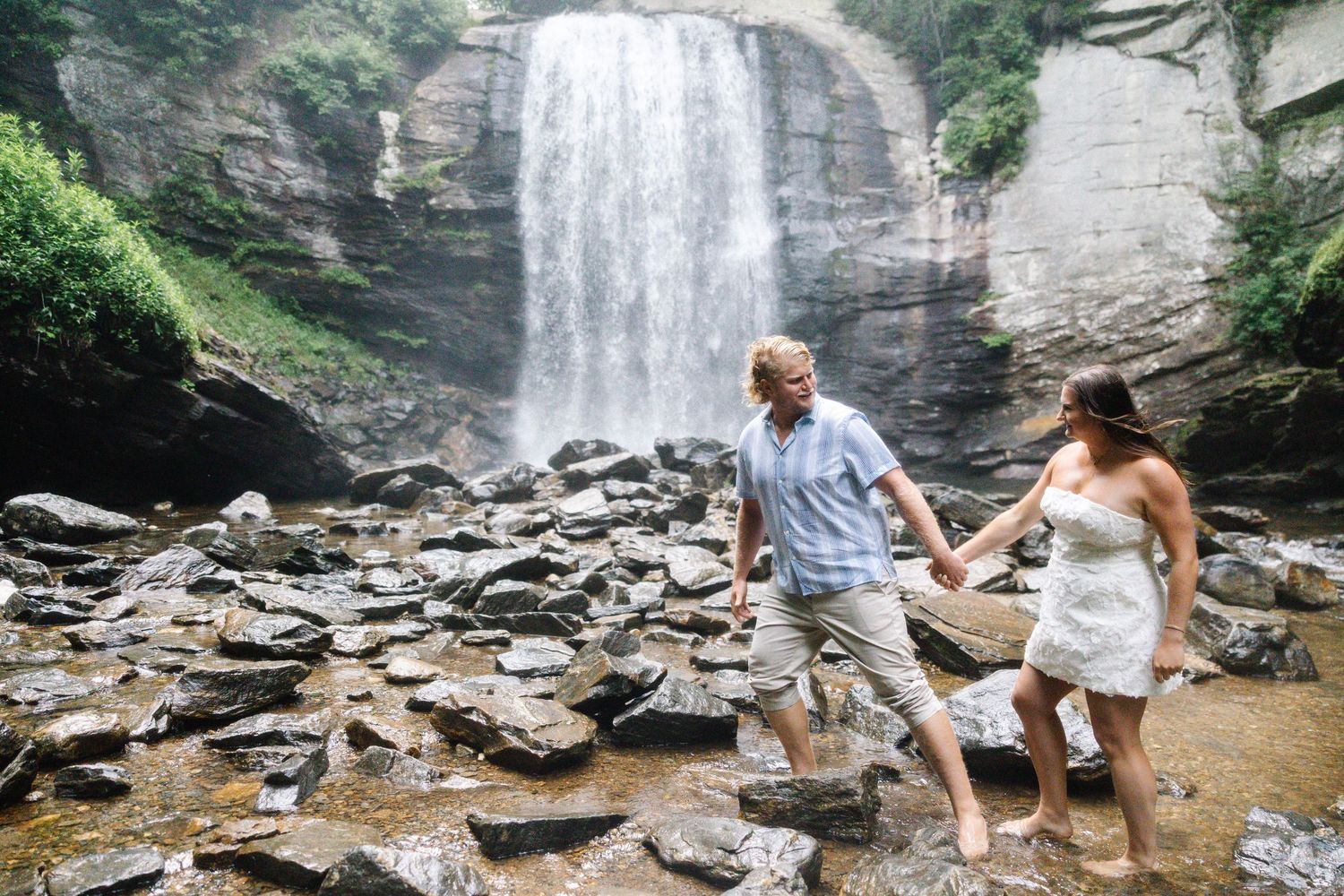 Couple holding hands while walking on rocks in front of a stunning waterfall in nature.